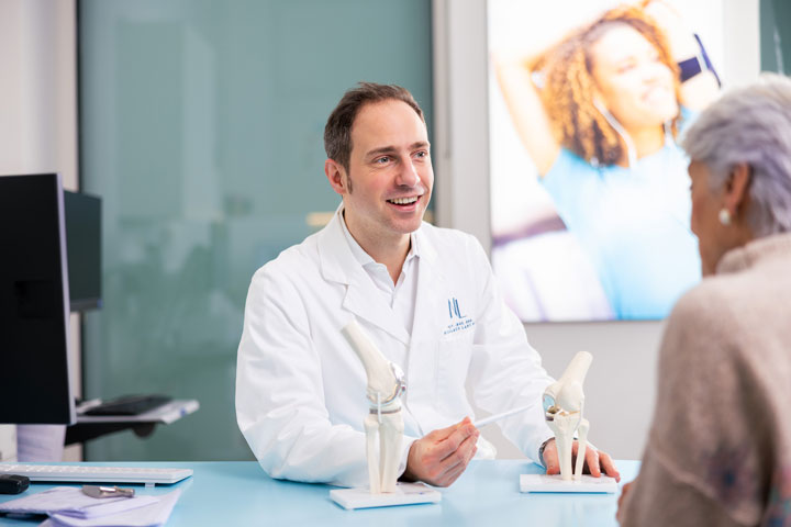 Dr. Nikolaus Lang berät Patientin in der Ordination, Knie-Modelle am Tisch.